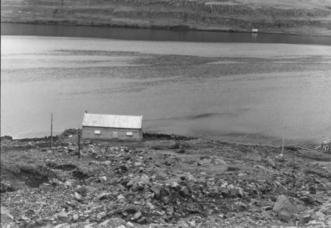 Ólafur Jónsson, ráðunautur og rithöfundur,  tók þessa mynd. Þetta er verbúð/hús sem stendur á aurkeilu Þófalækjar. Ólafur er þarna að skoða ummerki skriðunnar sem féll á hús sem stóð aðeins ofar við Þófalæk. Fimm manns létust þegar skriðan féll, 19. ágúst 1950. Talið er að myndin sé tekin nokkrum árum eftir skriðufallið, líklega 1954 eða 1955.