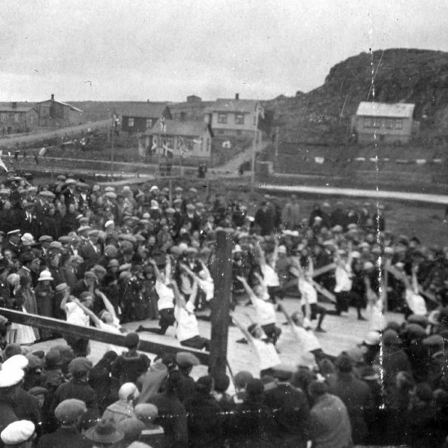 Hamarskotshamar í Hafnarfirði í fjarska. Ljósmyndarinn stendur á „skólamölinni“ við Barnaskóla Hafnarfjarðar, síðar Lækjarskóla, og leikfimiflokkurinn er á skólalóðinni. Gatan vinstra megin á myndinni er Öldugata. Langa húsið handan Öldugötu var oft kallað „bæjarbyggingin“. Lesandi, sem sendi upplýsingar, segir meðal annars: „Lengst til hægri á myndinni er hús sem mér sýnist vera með flatt þak og var að jafnaði kallað „kassahúsið“, einnig eftir að á það hafði verið sett venjulegt hallandi þak.“ Líklega er þetta Jónsmessuhátíð 29. júní 1924.  Minjasafnið á Akureyri / Mynd 29-358