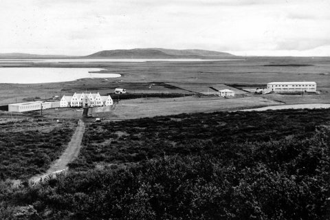 Myndin er frá Laugarvatni. Sigurður Kristinsson, prófessor við Háskólann á Akureyri, er alinn þar upp og sendi m.a. eftirfarandi: Myndin er tekin ofan úr fjallshlíðinni ofan við lundinn í kringum styttuna af Jónasi frá Hriflu, sem horfir niður eftir grasrennunni í skóginum á framhlið héraðsskólabyggingarinnar frá 1928, en Jónas var dóms- og kennslumálaráðherra 1927-1932. Til vinstri með sléttu þaki er viðbyggingin sem í var íþróttasalur og sundlaug, byggt 1942 en rifið á 10. áratugnum. Niðri á sléttlendinu fyrir aftan héraðsskólann er skólastjórabústaður Íþróttakennaraskólans, sem ég man eftir að hafa gengið undir nafninu Fuglinn. Hægra megin á myndinni er fyrst einbýlishúsið Garður, sem var lengi skólastjórabústaður Héraðsskólans en síðar bústaður skólameistara Menntaskólans. Lengst til hægri er svo elsti hluti húss Menntaskólans að Laugarvatni, byggður um og upp úr 1950. Við þessa byggingu var byggður turn á árunum 1959-60 þannig að myndin er tekin fyrir þann tíma. Myndin getur hins vegar ekki verið eldri en frá 1957 því að þá fyrst var lokið endurbyggingu Héraðsskólahússins eftir að burstirnar brunnu 1947.

Í baksýn eru Laugarvatn nær og Apavatn fjær. Aftan við Apavatn sést í Mosfell og þar fyrir aftan Vörðufell. Skógurinn í forgrunni er mun lægri á myndinni en hann er í dag, enda var þá fé beitt á skóginn og skógrækt ekki hafin. Minjasafnið á Akureyri / m-stae-1-5498 frá Minjasafninu á Akureyri