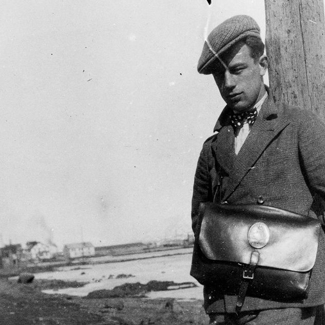 Þessi reffilegi maður mun vera Jón Pétursson sem fæddist á Akureyri 8. júní 1912 og lést 5. nóvember 2001. Jón var verkamaður og bifreiðastjóri í Vestmannaeyjum og síðar leigubílstjóri í Reykjavík. Á töskunni er gamla póstmerkið með kórónu og lúðri. Myndin er tekin í Fjörunni, í grennd við Nonnahús og Kirkjuhvol, þar sem Minjasafnið er nú til húsa. Stóra húsið næst á myndinni er Bæjarhúsið, þriggja hæða hús sem stóð þar sem gatan Duggufjara er núna.  Einnig sjást Tuliníusarhús og Bryggjuhúsið, steinsnar frá Höepfnersbryggjunni. / m-stae1-16388 frá Minjasafninu á Akureyri