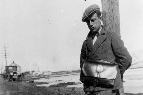 Þessi reffilegi maður mun vera Jón Pétursson sem fæddist á Akureyri 8. júní 1912 og lést 5. nóvember 2001. Jón var verkamaður og bifreiðastjóri í Vestmannaeyjum og síðar leigubílstjóri í Reykjavík. Á töskunni er gamla póstmerkið með kórónu og lúðri. Myndin er tekin í Fjörunni, í grennd við Nonnahús og Kirkjuhvol, þar sem Minjasafnið er nú til húsa. Stóra húsið næst á myndinni er Bæjarhúsið, þriggja hæða hús sem stóð þar sem gatan Duggufjara er núna.  Einnig sjást Tuliníusarhús og Bryggjuhúsið, steinsnar frá Höepfnersbryggjunni. / m-stae1-16388 frá Minjasafninu á Akureyri