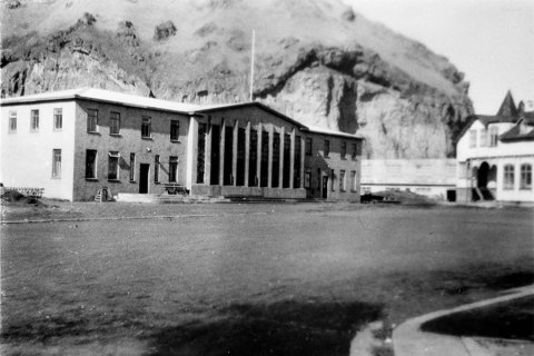 Þetta er í Vestmannaeyjum, Heimaklettur í baksýn. Eyjamaðurinn Guðni Einarsson (Gíslasonar í Betel), lengi blaðamaður á Morgunblaðinu, sendi: Þetta er Heimatorg á Heimaey. Til vinstri er Rafstöðin sem sá Eyjamönnum fyrir rafmagni með díselvélum og til hægri húsið Jómsborg. Svæðið og húsin fóru undir hraun í eldgosinu 1973. - / m-stae1-16309 frá Minjasafninu á Akureyri