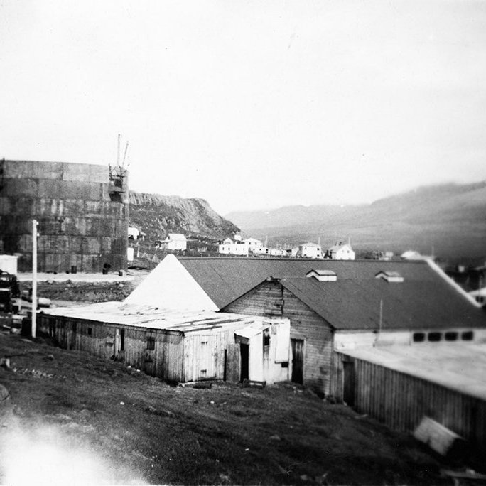 Þetta er á Skagaströnd. Myndin er tekin nálægt vestari enda Spákonufellshöfða nálægt svokölluðu Hólsnefi, skv. pósti frá Lárusi Ægi Guðmundssyni á Skagaströnd. Lengst til vinstri er lýsis- eða olíutankur fyrir síldarverksmiðju SR sem reist var á Skagaströnd á árunum 1943 – 1945, segir hann og á vef Héðins í Reykjavík kemur fram að um olíutank sé að ræða. Starfsmenn fyrirtækisins hafi lokið við að smíða tankinn árið 1947. Lárus Ægir segir: Byggingarnar neðst á myndinni eru nú allar horfnar en stóðu þar sem síðar var byggt frystihús en þar var jafnframt lítil bryggja sem byggð var ca. upp úr 1930. Sum íbúðarhúsanna sem sjást á myndinni eru enn á sínum stað en önnur eru horfin. - / m-stae1-15593 frá Minjasafninu á Akureyri