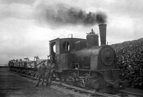 Myndin er tekin um 1925, í Reykjavíkurhöfn. Þetta er Eimreiðin Pionér í Austurhöfn, við hlið hennar standa eimreiðarstjórarnir Guðmundur Guðmundsson, til vinstri, og Páll Ásmundsson. Minør, systurlest Pionér, stendur á hafnarbakkanum við gömlu höfnina í Reykjavík á hverju sumri. Á vef Faxaflóahafna segir um þær: Eimreiðin Minør hefur ávallt verið í vörslu Faxaflóahafna yfir vetrartímann á meðan eimreiðin Pioner hefur verið varðveitt allt árið í kring á Árbæjarsafni. Eimreiðarnar voru keyptar hingað til lands vegna hafn­ar­gerðar.  Þar að auki var járn­braut lögð frá Öskju­hlíð,  Kringlumýri og Skóla­vörðuholt­inu./ m-stae-5-0417 frá Minjasafninu á Akureyri