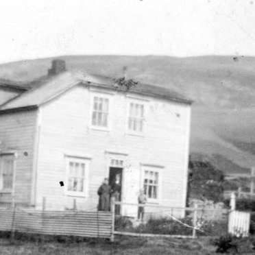 Þetta er gamli prestsbústaðurinn á Grenivík. Björn Ingólfsson, fyrrverandi skólastjóri á Grenivík, sendi fróðlegar og skemmtilegar upplýsingar, einu sinni sem oftar. Björn skrifar:
Húsið var byggt 1903 fyrir sóknarprestinn í Grenivík, séra Árna Jóhannesson. Hann bjó þar með stórri fjölskyldu til dauðadags 1927. Þá flutti í húsið Jóhann J. Kristjánsson læknir sem líka átti stóra fjölskyldu. Þau bjuggu þar til 1937 er þau fluttu til Ólafsfjarðar. Þá var húsið rifið og byggt nýtt læknishús. Símstöð var í húsinu frá 1919 til 1933.
Myndin er tekin eftir 1931 því að í kirkjugarðinum má sjá minnismerkið sem er á leiði sonar lækinshjónanna Hannesar Jóhannssonar (1926-1931). Líklega er þetta frú Inga Guðmundsdóttir sem stendur á pallinum - / m-stae1-17502 frá Minjasafninu á Akureyri