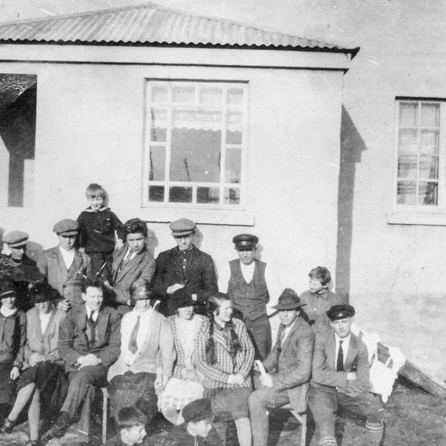 Myndin er tekin við Munkaþverá í Eyjafirði. Una Margrét Jónsdóttir, útvarpskona, skrifaði eftirfarandi: „Ég er nú búin að sýna Kristínu Jónsdóttur móður minni (Gígí) myndina. Það er rétt að hún er tekin á Munkaþverá sunnan undir húsveggnum. Samkvæmt áliti mömmu eru þessir á myndinni, talið frá vinstri. Aftari röð: Ingólfur Pálsson frá Uppsölum, óþekktur, Jónas Þorleifsson frá Grýtu (eða bróðir hans), svo Jónas Halldórsson frá Rifkelsstöðum. Drengurinn er líklega Jón Stefánsson á Munkaþverá, þá kemur óþekktur maður, svo Garðar Halldórsson frá Rifkelsstöðum og síðast Marinó Jónsson frá Uppsölum. (Fyrst var mamma reyndar á því að sá ysti til hægri væri Þorbjörn eða Sigurjón frá Borgarhóli, en hallaðist svo að því að það væri Marinó.) Þá kemur fremri röð frá vinstri: Líklega er Stefanía frá Steinsstöðum í Öxnadal yst t.v. þær næstu tvær álítur mamma að séu frænkur okkar, tvíburarnir Laufey og Sigríður Stefánsdætur, en er þó ekki alveg viss. Þá kemur Sigríður Halldórsdóttir frá Rifkelsstöðum, svo óþekktur maður og þrjár óþekktar konur, en ystur til hægri er, samkvæmt áliti mömmu, Valtýr Jónsson frá Borgarhóli. Allra fremst sjást höfuð tveggja ungra pilta og heldur mamma að sá sem er til vinstri geti verið Jóhann Rist. Þess skal líka getið að mömmu finnst líka koma til greina að stúlkan með flétturnar, þriðja frá hægri, sé Sigríður Stefánsdóttir, en þá getur hún náttúrlega ekki verið þriðja frá vinstri.“