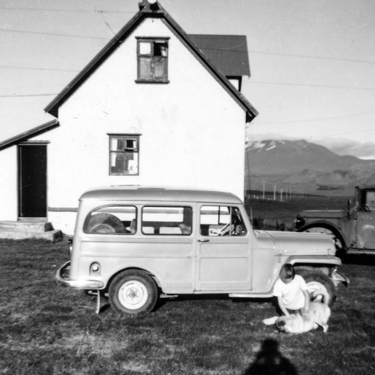 Myndin er tekin á bænum Leirubakka í Landsveit. Húsið á myndinni er „gamli bærinn sem búið er að rífa núna, ábúendur á þessum tíma voru Magnús Sigurðsson og Jóhanna Jónsdóttir,“ skrifaði Gunnar Sigurjónsson í skeyti til Akureyri.net. Magnús og Jóhanna voru afi hans og amma.

Stúlkan á myndinni er Jóhanna Sigurjónsdóttir, systir Gunnars. Honum sýndist það og Jóhanna staðfesti að svo sé. Hundurinn á myndinni hét Kópur, segir Jóhanna og bætir við að myndin sé tekin á tímabilinu 1963 til 1965.

Jóhanna segist hafa verið mörg sumur í sveit hjá afa sínum og ömmu á Leirubakka. Móðurbræður hennar áttu hins vegar bílana á myndinni; nær er Willys Overland árgerð 1951, segir Jóhanna, sem notaður var til að fara í réttir í Réttarnesi. „Bekkir voru sitt hvoru megin aftur í og þar var spjallað, sungið og teigaðar guðaveigar, þar smakkaði ég ginger ale í fyrsta sinn – var notað sem bland.“

Hinn bíllinn er Vibon, sennilega árgerð 1948 að sögn Jóhanna. Sá var notaður þegar fairð var í veiðiferðir í Veitivötn á Landmannaafrétti, að sögn hennar.