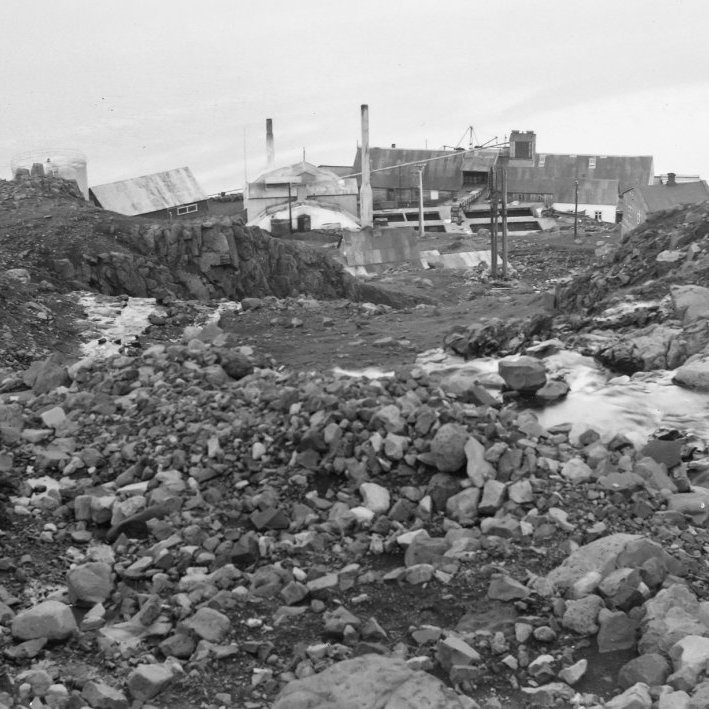 Þetta er á Seyðisfirði árið 1950. Myndin er tekin niður farveg Hæðarlækjar og í átt að húsunum Hæð og Ytri-Hæð og síðan gömlu síldarbræðslunni þar fyrir neðan.  Ólafur Jónsson ráðunautur og rithöfundur tók myndina. –Rannsóknarskýrsla um skriðuföll á Seyðisfirði sem kom út 2016 má finna á þessari slóð:
http://www.vedur.is/gogn/snjoflod/haettumat/se/haettumatskynning-agust-2019/2017_001_ISOR_Seydisfj_skridurannsoknir2016.pdf

Þar segir meðal annars: „Örlagaríkasta skriðufallið sem vitað er um á Seyðisfirði varð í Þófalæk þann 19. ágúst 1950. Þá braut skriða niður íbúðarhús að Strandarvegi 26 og varð fimm manns að bana.“ Fram kemur að Ólafur Jónsson fór á vettvang sumarið eftir og skoðaði öll ummerki vandlega. Önnur skriða hafði fallið nákvæmlega á sama tíma niður Hæðarlæk og valdið tjóni á Síldarverksmiðjunni.“