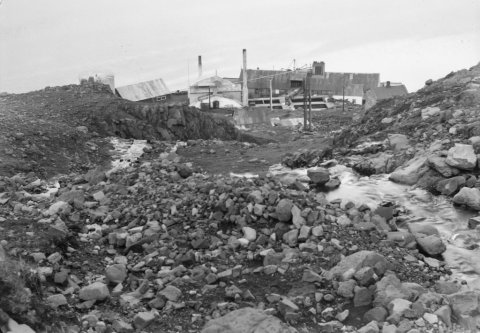 Þetta er á Seyðisfirði árið 1950. Myndin er tekin niður farveg Hæðarlækjar og í átt að húsunum Hæð og Ytri-Hæð og síðan gömlu síldarbræðslunni þar fyrir neðan.  Ólafur Jónsson ráðunautur og rithöfundur tók myndina. –Rannsóknarskýrsla um skriðuföll á Seyðisfirði sem kom út 2016 má finna á þessari slóð:
http://www.vedur.is/gogn/snjoflod/haettumat/se/haettumatskynning-agust-2019/2017_001_ISOR_Seydisfj_skridurannsoknir2016.pdf

Þar segir meðal annars: „Örlagaríkasta skriðufallið sem vitað er um á Seyðisfirði varð í Þófalæk þann 19. ágúst 1950. Þá braut skriða niður íbúðarhús að Strandarvegi 26 og varð fimm manns að bana.“ Fram kemur að Ólafur Jónsson fór á vettvang sumarið eftir og skoðaði öll ummerki vandlega. Önnur skriða hafði fallið nákvæmlega á sama tíma niður Hæðarlæk og valdið tjóni á Síldarverksmiðjunni.“